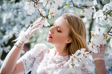 portrait of beautiful blonde smiling romantic bride in a white dress walking in blossoming magnolia...