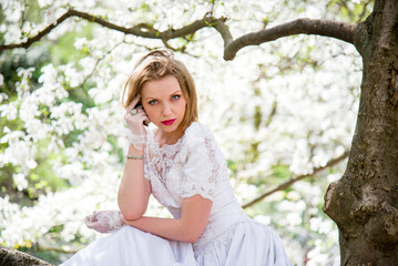 beautiful blonde smiling romantic bride in a white dress sitting on a branch in blossoming magnolia garden on sunny spring day