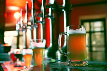 Glass, beer mug filled with golden-colored lager or pilsner foamy beer. Blurred background of bar, pub atmosphere. Concept of beer drink, alcohol, brewery, pub atmosphere, taste - obrazy, fototapety, plakaty