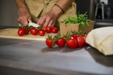 Close-up view of a male chef's hands chopping tomato cherry on a cutting board. Ingredients on the kitchen table. Man cooking healthy salad in the minimalist home kitchen interior