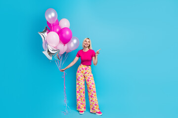 Full body photo of attractive young woman hold air balloon point empty space dressed stylish pink clothes isolated on blue color background