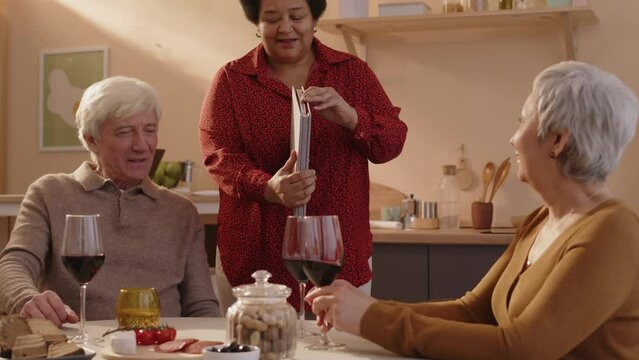Medium shot of happy senior Black woman showing photo album to her multiethnic old friends while having small dinner party in bright apartment