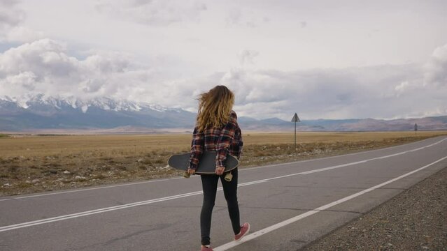 Woman holding a skateboard embark on a road trip, walking along an open highway towards distant, snow-capped mountains, embodying a sense of adventure and freedom. A carefree free girl walks with a