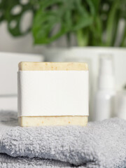 Soap bar with blank label on light grey towel against monstera plant in bath, mockup