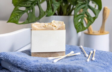 Soap bar with blank label on blue towels near basin and green monstera in bath, mockup