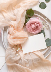 Card near cream fabric knot and light pink roses on plates top view copy space, wedding mockup
