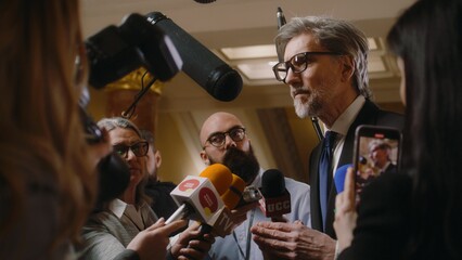 Politician answers press questions and gives interview for television news. Mature diplomat surrounded by crowd of journalists in parliament hall. Press conference. Inspirational speech. Slow motion. - obrazy, fototapety, plakaty