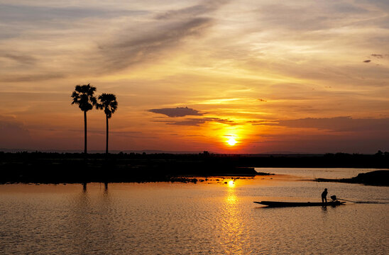 Silhouette of fisherman on boat with two palm trees in the lake at sunset.