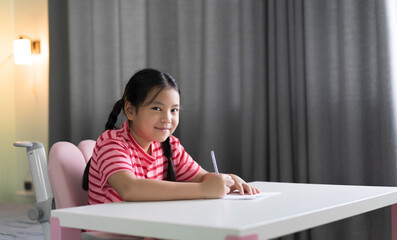 learn from home by asian child or kid girl student smile writing on paper to draw learning studying and fun doing homework in classroom homeschool and bedroom with warm white lamp and window light