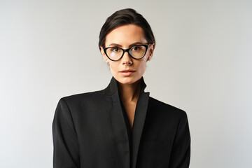 A stylish woman wearing glasses and a black jacket poses against a grey studio background.