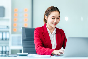 Asian young businesswoman, a Financial Advisor, works happily in her office. She uses her laptop and mobile phone to analyze charts and communicate financial strategies. Red formal suit