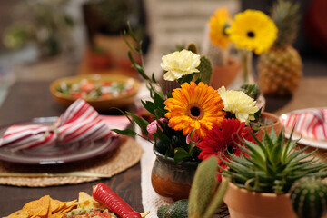 Obraz na płótnie Canvas Bunch of gerberas and carnations in vase standing in the center of festive table next to domestic flowers in flowerpots and homemade snacks