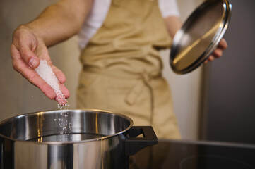Close-up of a chef adding salt into a boiling water in the stainless steel saucepan, standing at an electric stove, boiling water for spaghetti or other meal