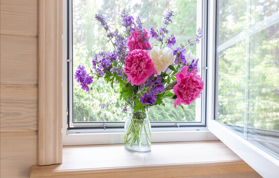 Open white window with a mosquito net in a rustic wooden house. Summer bouquet of peonies on the windowsill
