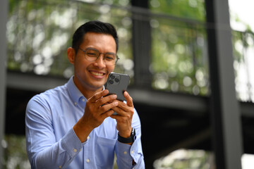 Smiling businessman in eyeglasses texting on mobile phone sitting on the stairs outside