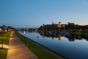 Krakow, Poland. 09.10.2022 Historic Wawel Castle in the evening blue hour on the banks of the Vistula river and floating ships restaurants.