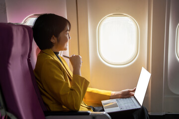 Smiling businesswoman in a yellow blazer using a laptop on a tray table, working efficiently on an airplane.
