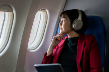 A thoughtful woman wearing a red jacket and headphones looks out an airplane window while holding a tablet.