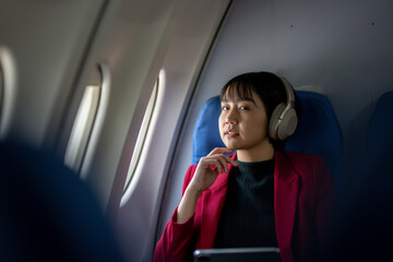 A thoughtful woman wearing a red jacket and headphones looks out an airplane window while holding a tablet.