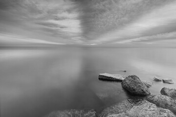 Black and white photo of ominous striped clouds above a calm Markermeer with stones in the...