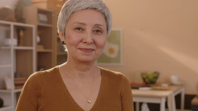 Tilt up portrait of mid-aged Asian woman revoking memories by looking at photos in photo album then smiling at camera, standing in bright cozy apartment with sun rays going through window
