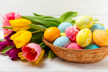 Easter basket filled with colorful eggs and a bouquet of tulips on a textured wooden table. Easter celebration concept. Colorful easter handmade decorated Easter eggs. Place for text. Copy space