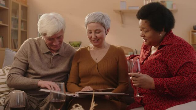 Medium shot of three diverse senior friends sitting on couch in bright cozy living room, talking and looking at photos in photo album, reminiscing and revoking memories