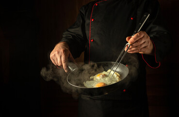 Cropped silhouette of a chef with a hot frying pan for cooking scrambled eggs for breakfast. Concept on a dark background of frying an egg