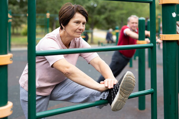 Aged woman doing stretching on sports bars in open-air sports area