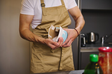Close-up man chef in beige apron, pouring some salt from paper bag, seasoning meal while cooking dinner