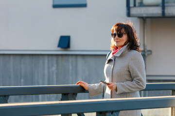 Happy mature woman enjoying a sunny morning walk on a bridge, dressed in casual chic attire with a...