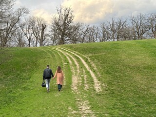 A guy and a girl are walking along a country road. A man and a woman hold hands and walk along the path, rear view. A couple in love climb a mountain, walk along the hill.