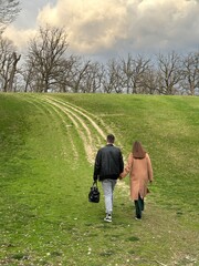 A guy and a girl are walking along a country road. A man and a woman hold hands and walk along the path, rear view. A couple in love climb a mountain, walk along the hill.