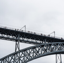 Tourists walking, taking photos and a woman posing from the upper platform of the Don Luis I steel bridge where the Porto metro passes with rain clouds.