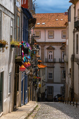 Steep downhill cobblestone street with balconies of old houses in a classic and old neighborhood of Porto, decorated with colored paper flags and lanterns, flowers, pots and antennas. Portugal.