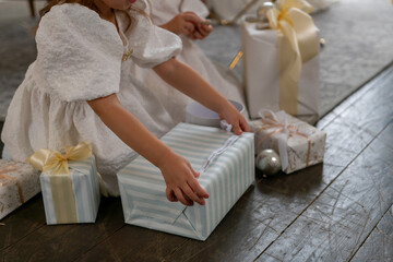 A young girl is opening a gift box with a ribbon on it. The scene is set in a room with a few other...
