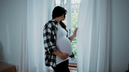 Contemplative pregnant woman standing by window curtains gazing outside from residence home,...