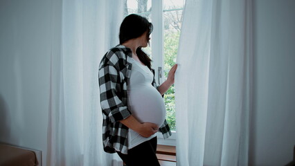 Contemplative pregnant woman standing by window curtains gazing outside from residence home, maternity scene, 8 months belly