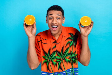 Photo of excited funky guy dressed print shirt holding two orange halves isolated blue color...