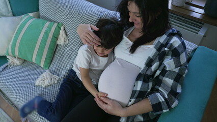 Tender child hugging pregnant mother in display of affection to unborn baby brother seated on couch...