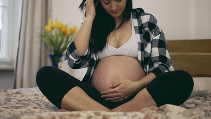 Third trimester pregnancy - woman caresses pregnant belly in 8 month expecting baby seated in bed...