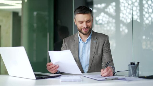Busy businessman working fills out documents, writing while sitting at desk at workplace in business office. Confident man financier or accountant is doing paperwork accounting or making a tax return
