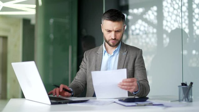 Serious busy businessman is doing paperwork filling out a form on laptop computer sitting at workplace in business office. Focused thinking man financier or accountant working making financial report