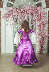 Young beautiful woman in fantasy rococo style medieval dress standing near wall with pink flowers back pose