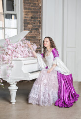 Beautiful woman in fantasy white and purple rococo style medieval dress sitting near piano with pink flowers