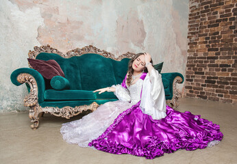 Beautiful sad crying woman in fantasy white and purple rococo style medieval dress sitting on the floor near sofa