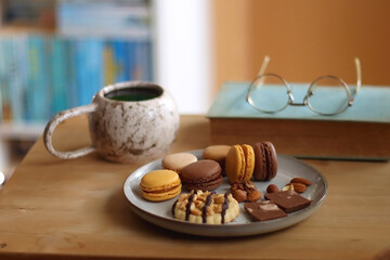 Plate with macarons, cookies, chocolate and nuts, cup of tea or coffee, book and reading glasses on the table. Selective focus. Colorful bookcase in the background.