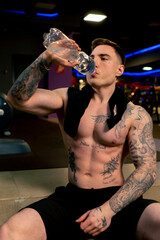 in the gym young handsome guy with tattoos sitting drinking water from a plastic bottle resting
