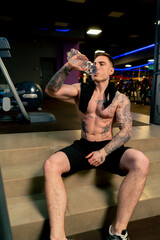 in the gym young handsome guy with tattoos sitting drinking water from a plastic bottle resting