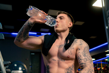 close up in the gym young handsome guy with tattoos sitting drinking water from a plastic bottle...